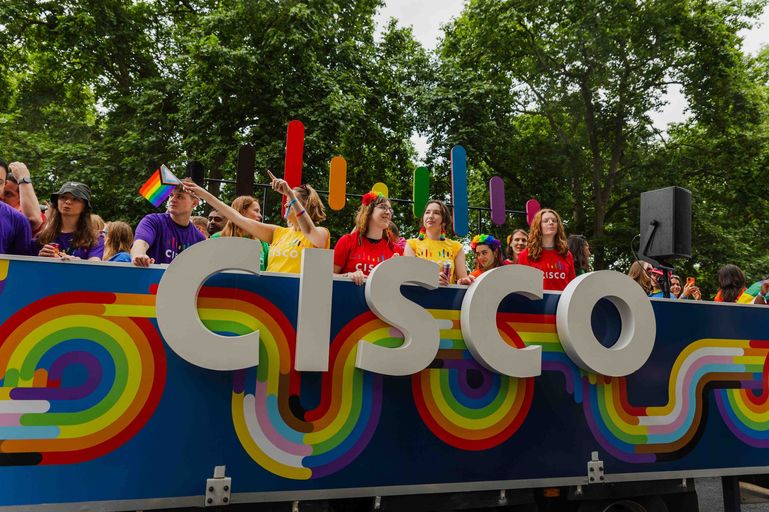 A colorful Pride parade float branded with a large “CISCO” logo features rainbow-themed graphics and tall, multicolored bars resembling a stylized signal icon. People on the float wear Cisco shirts in red, yellow, and purple and wave Pride flags while smiling and engaging with the crowd under a canopy of green trees during London Pride Parade 2025.