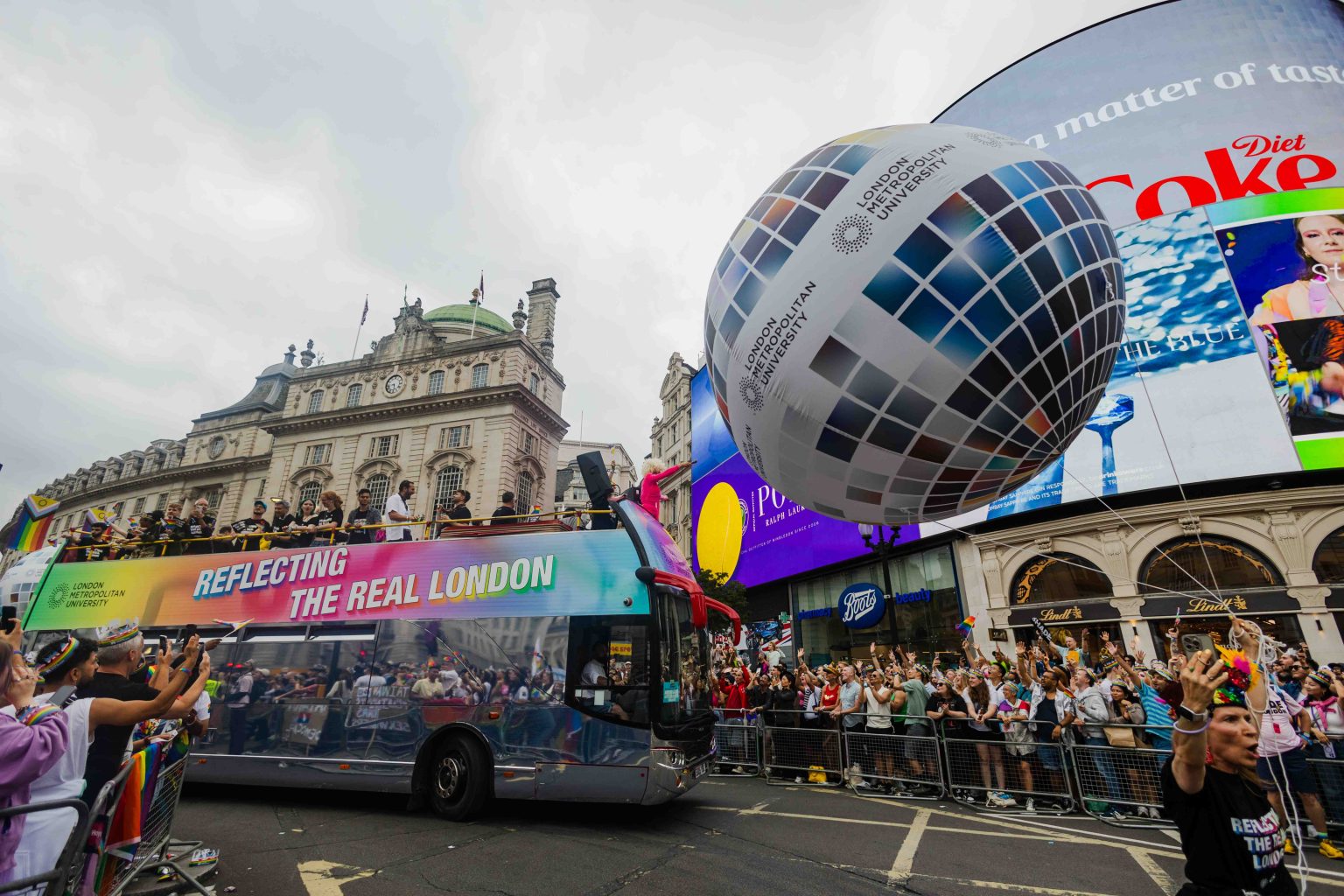 A colorful double-decker bus from London Metropolitan University with the slogan “Reflecting the Real London” rolls through a crowded Pride parade 2025 at Piccadilly Circus. A large balloon resembling a mirrored disco ball, also branded with the university’s name, floats above cheering spectators lined along barricades.