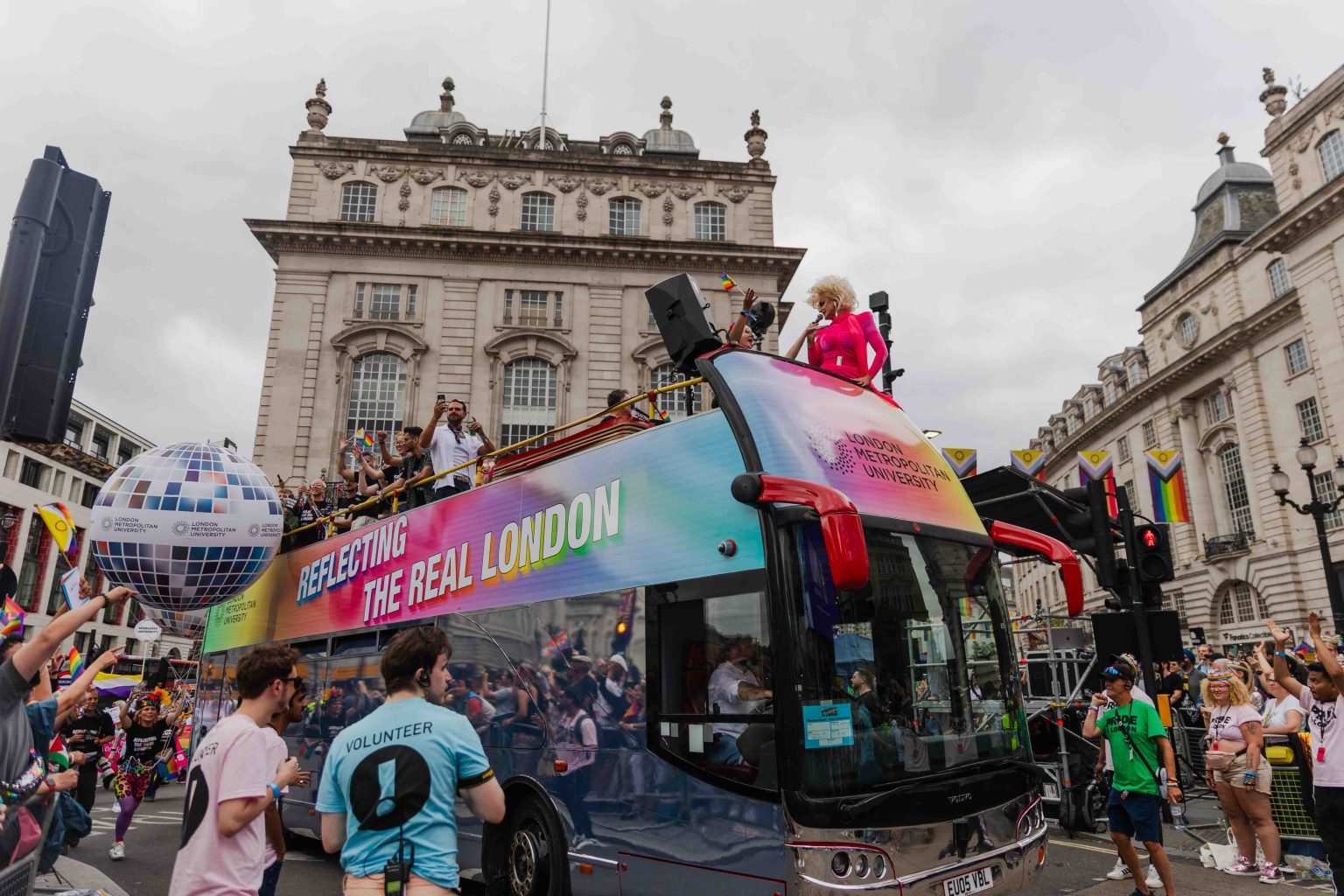 A London Metropolitan University double-decker bus (their pride parade float vehicle for London pride 2025) with a vibrant rainbow gradient and the slogan “Reflecting the Real London” moves through the Pride parade, with a drag performer in bright pink standing at the front. Spectators cheer alongside a large disco ball balloon bearing the university’s logo, while volunteers and parade-goers wave flags and capture the moment.