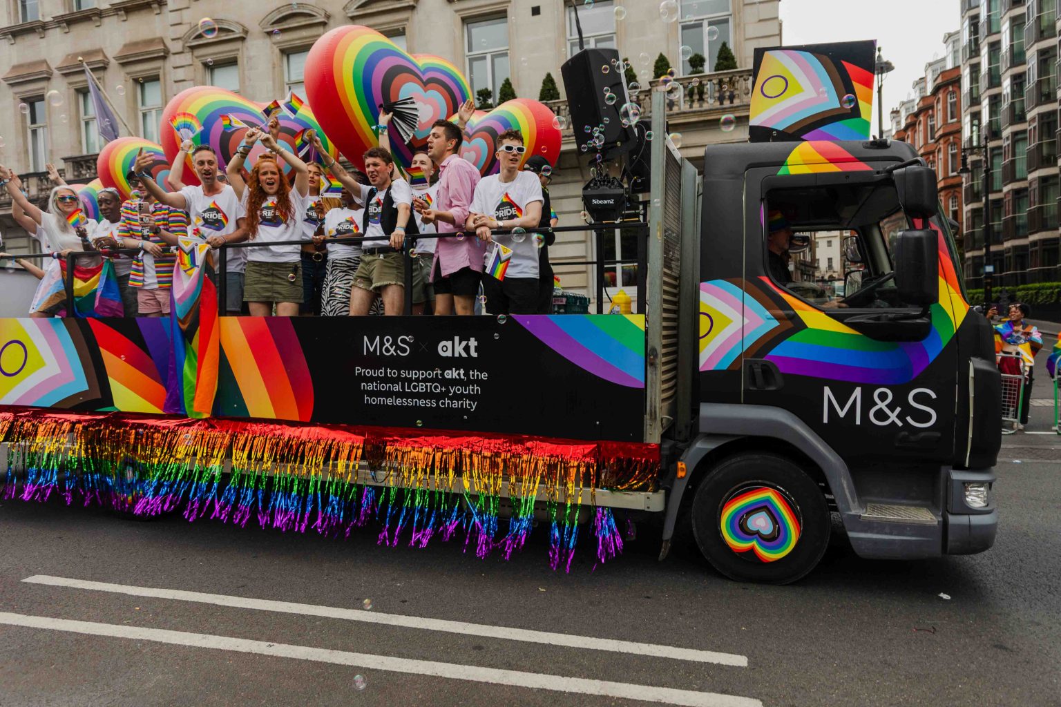 A colorful M&S Pride parade float for London Pride 2025 features rainbow fringe, oversized heart decorations, and a banner supporting akt, a national LGBTQ+ youth homelessness charity. People on the float cheer and wave Pride flags, all wearing “M&S Pride” shirts against a backdrop of festive rainbow colors.