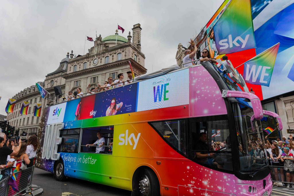 A colorful double-decker Sky bus decorated in a rainbow gradient with pink sparkles passes through central London during Pride parade 2025. The side displays digital screens with the words “WE” and “believe in better” while people onboard wave Sky-branded rainbow flags above a cheering crowd.