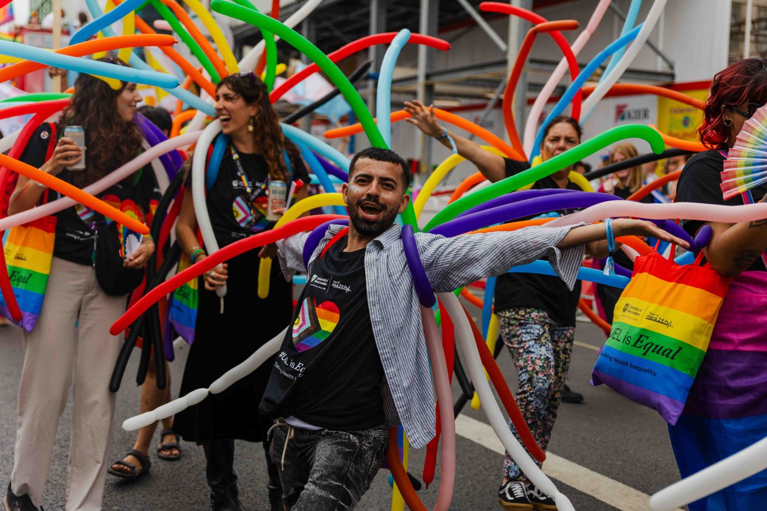 A lively group of Pride parade participants wear black shirts with a rainbow heart logo and are covered in long, colorful balloon tubes extending outward like streamers. One person in front poses joyfully with arms wide open, while others laugh and interact behind them