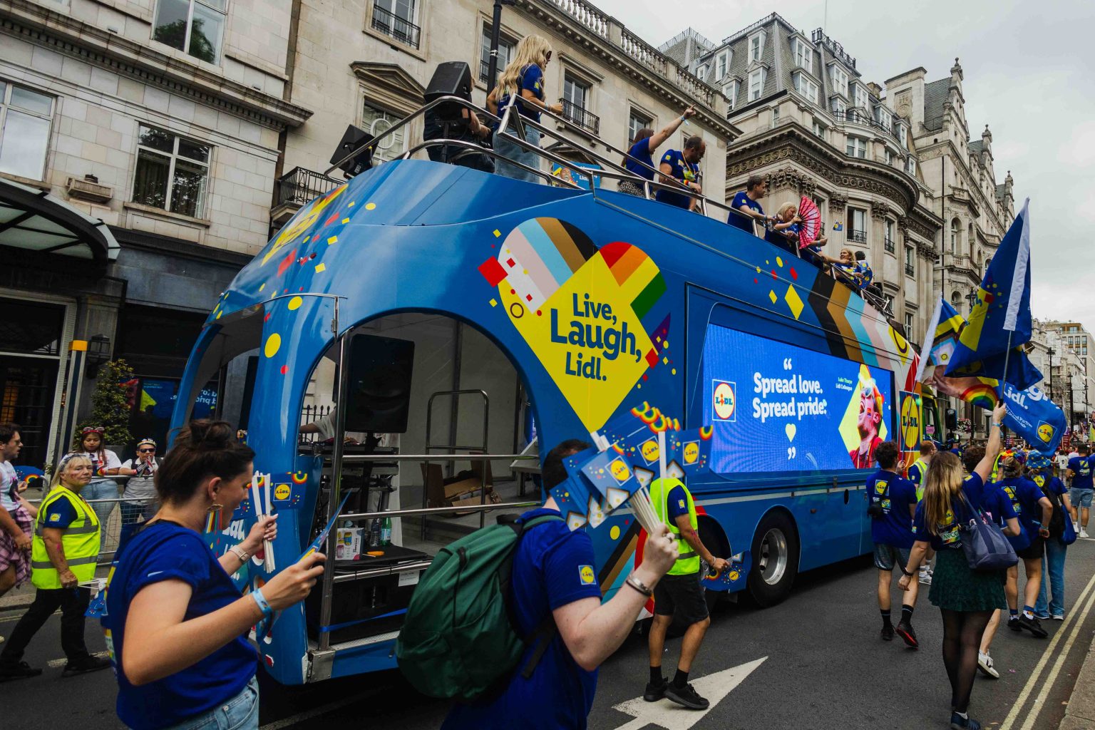 A bright blue Lidl Pride parade bus features a rainbow heart graphic with the slogan “Live, Laugh, Lidl” and a large digital screen displaying “Spread love. Spread pride.” Participants in matching blue shirts walk alongside, waving Lidl-branded fans and flags while engaging with the crowd on a busy city street. Side angle of lidl's london pride parade 2025 vehicle.
