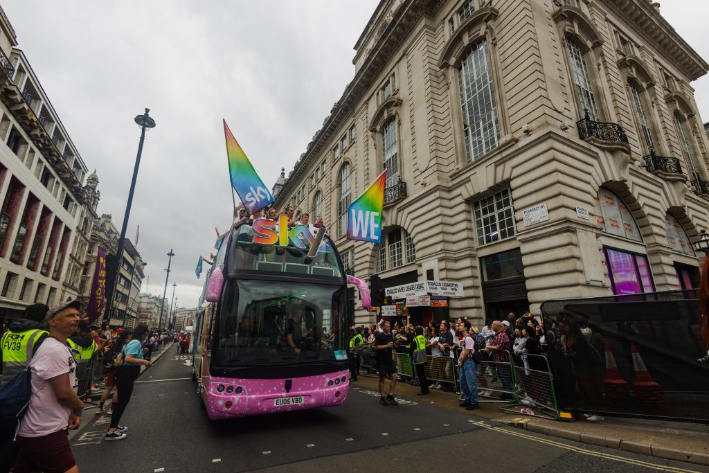 LED HyperBus promotional bus moving through central London Pride parade with Sky branding surrounded by crowds and Piccadilly Circus buildings