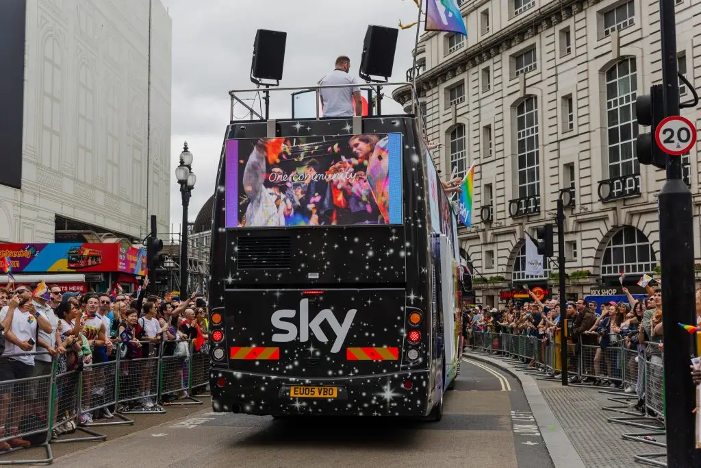 LED HyperBus promotional bus back view approaching through London street during Pride with Sky LED content and large crowds behind barriers
