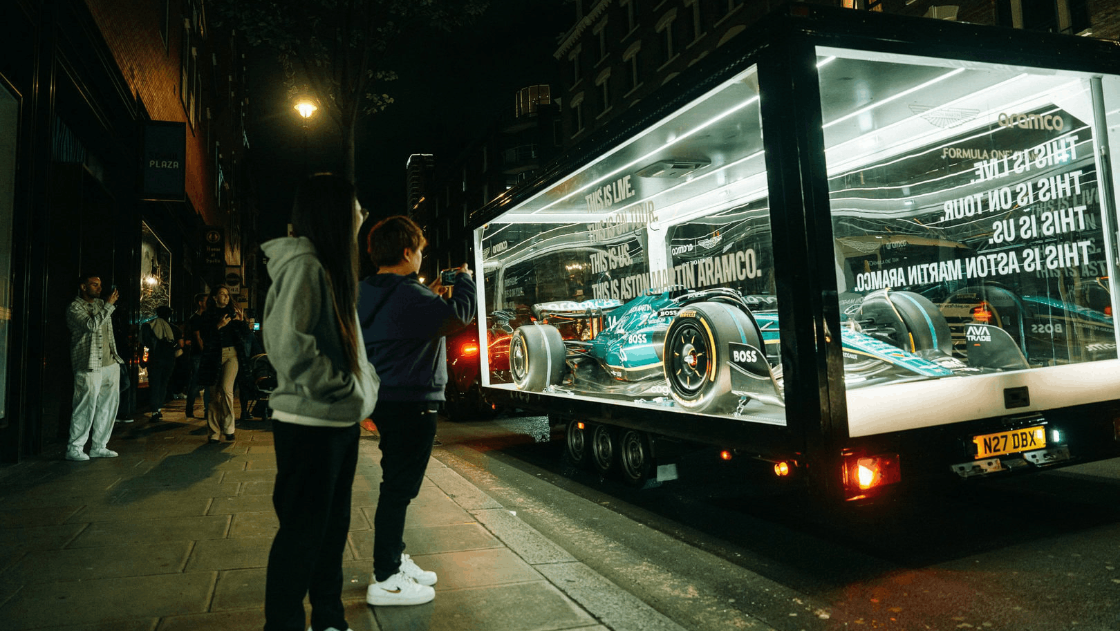 Aston Martin Aramco F1 car illuminated at night on a London street inside The Clear Idea Transparent Showcase Trailer with integrated lighting