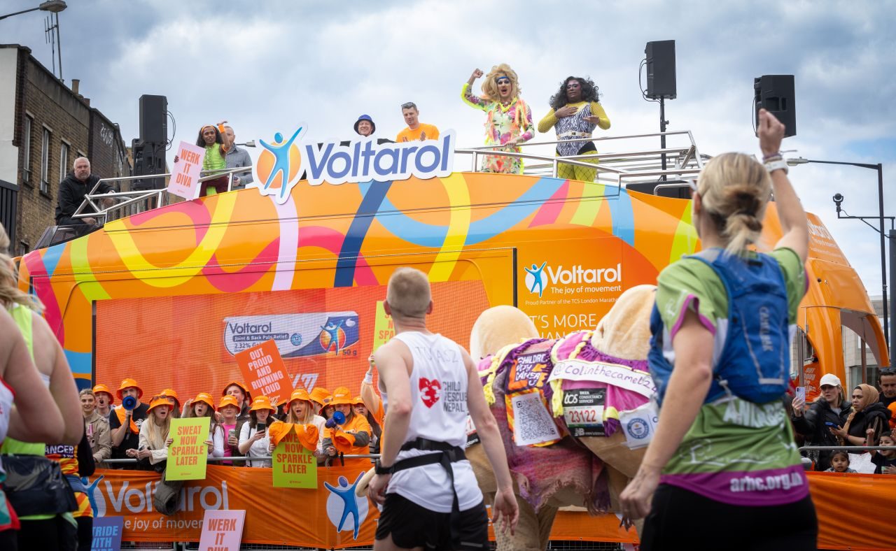 ShowTruck with Voltarol branding at marathon cheering point with performers on upper deck engaging crowd and runners passing in foreground